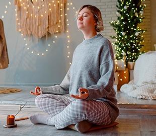 Mulher em pose de lótus, meditando com um incenso aceso ao lado, em um ambiente decorado com luzes de Natal e uma árvore, simbolizando bem-estar e relaxamento na época natalina.
