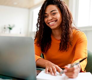 Mulher sorridente com cabelo cacheado, usando um laptop e anotando em um caderno, simbolizando a facilidade e praticidade de uma central de serviços online.
