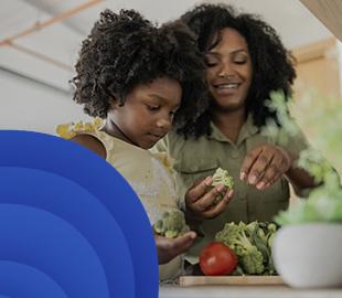 Mãe e filha sorrindo, preparando brócolis e outros vegetais na cozinha, simbolizando a importância de cuidar da alimentação com a ajuda de uma teleconsulta nutricional.