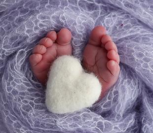 Pés de um recém-nascido envoltos em um cobertor lilás, com um coração branco e fofo de feltro sobre eles, simbolizando o Novembro Roxo e a prevenção da prematuridade.