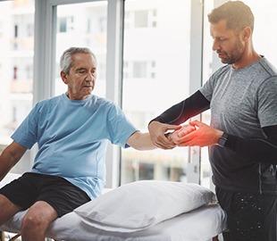 Fisioterapeuta examinando o punho de um paciente idoso, com uma área vermelha destacada indicando dor, simbolizando a ortopedia, prevenção e cuidados essenciais.