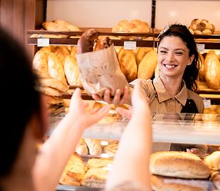 Mulher sorridente entregando um pão a um cliente em uma padaria, simbolizando o atendimento ao cliente e o funcionamento de pequenas empresas.