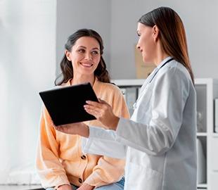 Médica de jaleco branco mostrando um tablet para uma paciente sorridente em um consultório, simbolizando a consulta e informações sobre plano de saúde.