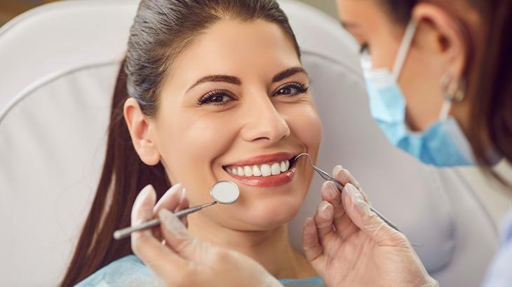 Mulher sorridente em consulta odontológica, com uma dentista usando máscara examinando seus dentes com espelho e sonda, ilustrando os benefícios de um plano odontológico.