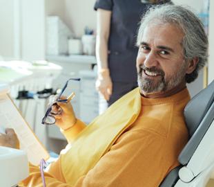 Homem sorridente sentado na cadeira do dentista, segurando óculos e uma prancheta, com um profissional de saúde ao fundo, ilustrando a consulta odontológica e a prevenção de doenças bucais.