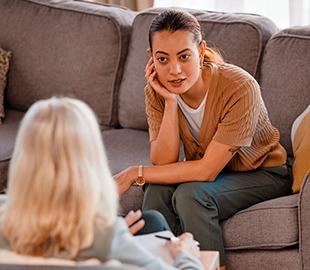 Mulher jovem atenta em uma sessão de terapia, olhando para a terapeuta sentada de costas, simbolizando atendimento psicológico e saúde mental.