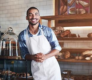 Jovem empreendedor, usando um avental, sorrindo e com as mãos cruzadas em sua padaria ou confeitaria, simbolizando a importância do plano de saúde para PMEs.