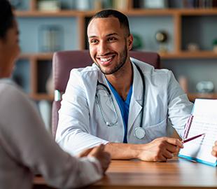 Médico sorridente de jaleco branco, com estetoscópio, conversando com um paciente em um consultório, simbolizando a comunicação de um aviso importante, como a reforma de uma clínica.