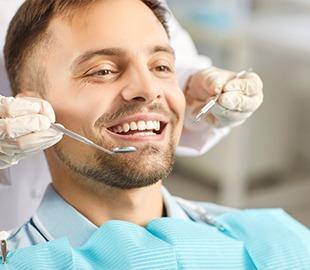 Homem sorrindo feliz enquanto um dentista com luvas examina seus dentes com instrumentos odontológicos, ilustrando os benefícios de um plano odontológico e saúde bucal.
