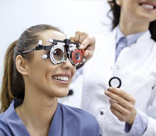 Mulher sorridente realizando um exame de vista com um foróptero, enquanto uma oftalmologista ajusta o aparelho, simbolizando os cuidados e exames para a saúde ocular.