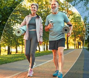 Casal idoso sorridente caminhando ao ar livre, segurando tapetes de yoga e garrafas de água, simbolizando a importância de hábitos saudáveis e ativos na terceira idade.
