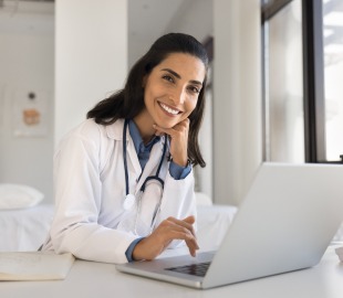 Médica sorridente usando jaleco e estetoscópio trabalha no notebook em consultório, representando atendimento humanizado e tecnologia na saúde