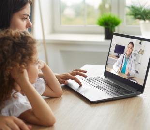 Mãe e filha pequena em frente a um notebook, realizando uma consulta de telemedicina pediátrica com uma médica na tela, simbolizando o atendimento online para crianças.