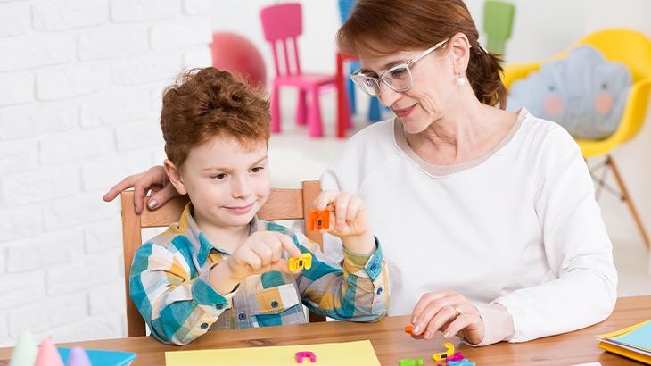 Mulher mais velha com óculos e um menino ruivo montando letras de brinquedo em uma mesa, simbolizando terapia educacional ou apoio ao aprendizado infantil.