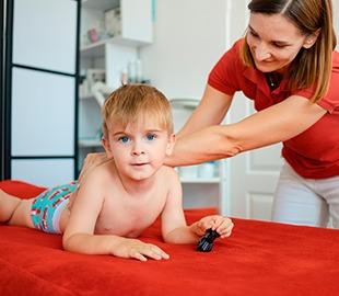 Criança deitada de bruços em uma maca vermelha, olhando para a câmera, enquanto uma terapeuta ocupacional o auxilia por trás, ilustrando a terapia ocupacional infantil.