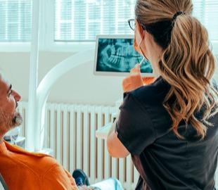 Dentista de óculos e cabelo preso mostrando um raio-X dental em um tablet para um paciente masculino, ilustrando a inclusão de dependentes em um plano odontológico.