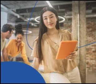 Mulher asiática sorridente, segurando um tablet, com colegas de trabalho desfocados ao fundo, simbolizando a análise de indicadores econômicos.