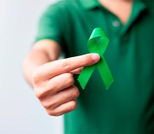 Mão segurando uma fita verde de conscientização, com uma pessoa vestindo uma camisa polo verde ao fundo, simbolizando a campanha Julho Verde e a conscientização sobre o câncer de cabeça e pescoço.