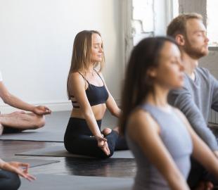 Grupo de pessoas meditando em pose de lótus em tapetes de yoga, com olhos fechados e expressão de tranquilidade, simbolizando os benefícios da meditação para o corpo e a mente.