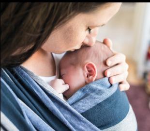 Mãe com cabelo escuro beijando a testa de seu bebê recém-nascido, que está em um sling de bebê, simbolizando o período pós-parto e o vínculo materno.