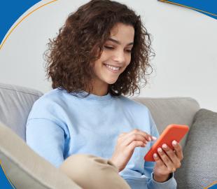 Mulher sorridente com cabelo cacheado, sentada em um sofá e usando o celular, simbolizando as mudanças e a facilidade de acesso à Ouvidoria Clinipam.