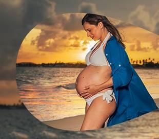 Mulher grávida em biquíni branco e camisa azul, com as mãos na barriga, posando em uma praia ao pôr do sol, simbolizando uma campanha institucional de saúde e maternidade.