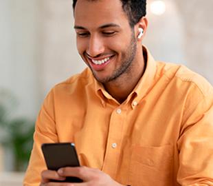 Homem sorridente de camisa laranja, usando fones de ouvido sem fio, olhando para o celular, simbolizando a nova experiência e facilidade de usar um plano de saúde através de dispositivos móveis.