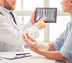 Médico de jaleco branco mostrando uma imagem de raio-X de ossos em um tablet para uma paciente com braço engessado, simbolizando o diagnóstico e tratamento da osteoporose e fraturas ósseas.