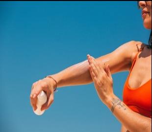 Mulher de biquíni laranja aplicando protetor solar no braço sob um céu azul, simbolizando os cuidados com a pele saudável no verão.