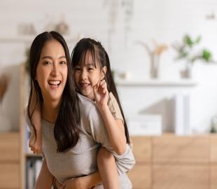Mãe e filha asiáticas sorrindo e se abraçando, com a criança nas costas da mãe, simbolizando alegria e a importância de escolher um plano de saúde para crianças.