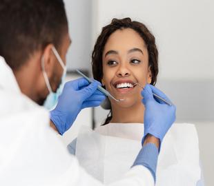 Mulher jovem sorridente na cadeira do dentista, enquanto um dentista com máscara e luvas azuis realiza um exame bucal com instrumentos, ilustrando um plano de tratamento odontológico e saúde bucal.