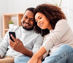 Casal feliz e sorridente, compartilhando fones de ouvido e olhando para um smartphone, simbolizando a comunicação e o acompanhamento de informações sobre o reajuste de planos de saúde.