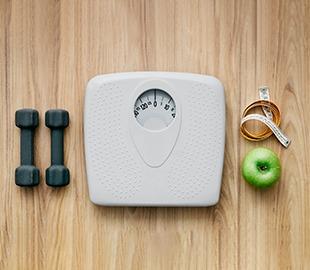 Balança, halteres, fita métrica e maçã verde sobre uma superfície de madeira, simbolizando a obesidade como problema de saúde pública, peso, exercícios e alimentação saudável.