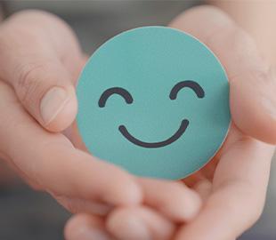 Mãos de um adulto segurando uma forma redonda verde-azulada com um rosto sorridente, simbolizando o cuidado e a saúde mental infantil.