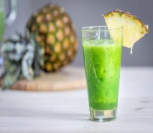 Copo de suco verde de couve com uma fatia de abacaxi na borda, e um abacaxi inteiro desfocado ao fundo, ilustrando os benefícios do suco de couve.