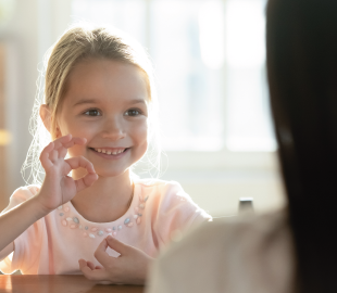 Menina sorrindo e fazendo o sinal de ok com as mãos enquanto interage com um terapeuta (fora de foco), simbolizando a ampliação da rede de prestadores de terapias e terapias especiais.