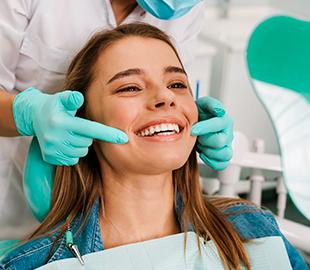 Mulher jovem sorridente na cadeira do dentista, enquanto um profissional com luvas azuis aponta para seu dente, simbolizando as vantagens de um plano odontológico.